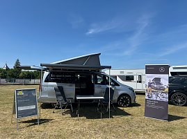 Ein silberner Mercedes-Benz Campingbus mit geöffnetem Aufstelldach und Markise steht auf einer Wiese beim Leichtathletik-Meeting Anhalt 2025. Vor dem Fahrzeug stehen ein Tisch mit Stühlen, ein Aufsteller und ein Werbebanner, im Hintergrund sind weitere Camper und Fahrzeuge zu sehen.