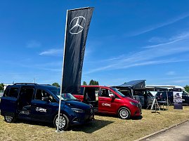 Mehrere Mercedes-Benz Fahrzeuge des Autohauses Peter, darunter ein blauer Van, ein roter Van und ein silberner Campingbus mit geöffnetem Aufstelldach, stehen auf einer Ausstellungsfläche beim Anhalt 2025 Leichtathletik-Meeting. Vor den Fahrzeugen weht eine schwarze Fahne mit Mercedes-Stern, rechts stehen Infotafeln. Der Himmel ist klar und blau.