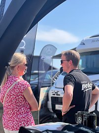 Ein Mitarbeiter des Autohauses Peter spricht auf dem Anhalt 2025 Leichtathletik-Meeting mit einer Besucherin. Im Hintergrund stehen mehrere Mercedes-Benz Fahrzeuge sowie Fahnen und ein Wohnmobil. Die Szene findet unter freiem Himmel bei sonnigem Wetter statt.