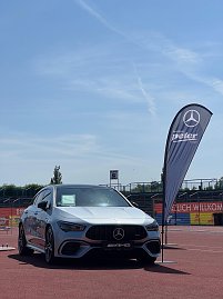 Ein silberner Mercedes-AMG steht auf der Laufbahn des Anhalt 2025 Leichtathletik-Meetings. Rechts neben dem Fahrzeug weht eine schwarze Fahne des Autohauses Peter im Wind. Im Hintergrund sind Tribünen und Werbebanner zu sehen, während der Himmel klar und sonnig ist.
