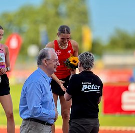Bei der Siegerehrung des Anhalt 2025 Leichtathletik-Meetings überreicht eine Mitarbeiterin des Autohauses Peter einer Athletin mit rotem Trikot und Sonnenblumenstrauß die Auszeichnung. Daneben steht ein älterer Mann im blauen Hemd, während im Hintergrund die Laufbahn und farbige Werbebanner sichtbar sind.