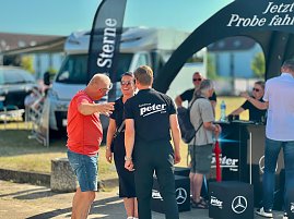 Menschen unterhalten sich gut gelaunt am Stand des Autohauses Peter beim Anhalt 2025 Leichtathletik-Meeting. Im Vordergrund stehen zwei Personen in schwarzer Autohaus-Peter-Kleidung und ein Mann im roten T-Shirt. Im Hintergrund sind weitere Besucher, ein Wohnmobil und schwarze Promotion-Zelte mit Mercedes-Benz-Logo zu sehen.