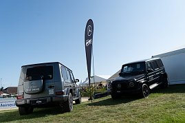 Zwei Mercedes-Benz G-Klasse Fahrzeuge stehen auf einer leicht geneigten Wiese neben wei&szlig;en Eventzelten. Ein graues Modell mit der Aufschrift �Electric Test Drive� ist von hinten zu sehen, daneben ein schwarzes Modell von vorne. Eine schwarze Fahne mit Mercedes-Logo steht zwischen den Fahrzeugen. [Mercedes-Benz G 580 mit EQ Technologie | Energieverbrauch kombiniert: 30,3-;27,7 kWh/100 km | CO2-Emissionen kombiniert: 0 g/km | CO2-Klasse: A] [Mercedes-Benz G 450 d | Energieverbrauch kombiniert: 9,9-8,7 l/100 km | CO2-Emissionen kombiniert: 261-227 g/km | CO2-Klasse: G]