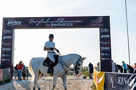 Ein Reiter in wei&szlig;em T-Shirt und schwarzem Helm sitzt auf einem wei&szlig;en Pferd unter einem Begr&uuml;&szlig;ungsbogensorenn mit der Aufschrift �Herzlich willkommen in Immenrode�. Der Bogen ist mit Logos verschiedener Spo versehen. Im Hintergrund sind mehrere Personen und ein sonniger Himmel zu sehen.