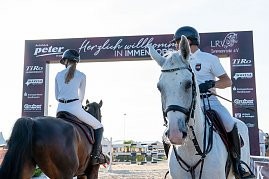 Zwei Reiter in wei&szlig;er Turnierkleidung sitzen auf einem wei&szlig;en und einem braunen Pferd vor einem Begr&uuml;&szlig;ungsbogen mit der Aufschrift �Herzlich willkommen in Immenrode�. Im Hintergrund sind Reitplatz und Turnierkulisse zu sehen.