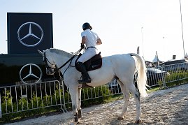 Ein Reiter in wei&szlig;er Turnierkleidung sitzt auf einem wei&szlig;en Pferd vor einem Mercedes-Benz Werbebanner mit zwei gro&szlig;en Logos. Der Reitplatz ist von Absperrgittern und gr&uuml;nen Str&auml;uchern umgeben, im Hintergrund stehen Fahrzeuge und Eventzelte.