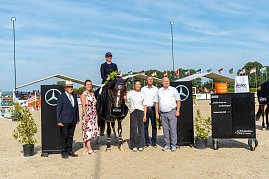 Siegerehrung beim Reitturnier in Immenrode: Ein Reiter sitzt auf seinem Pferd mit Siegerdecke und Blumenstrau&szlig;, daneben stehen f&uuml;nf Erwachsene zur Gratulation. Die Szene spielt sich zwischen Mercedes-Benz-Hindernissen und unter wehenden Fahnen auf dem Turnierplatz ab.