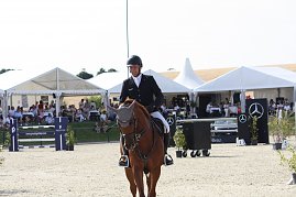 Ein Reiter in schwarzer Turnierjacke und Helm reitet auf einem braunen Pferd &uuml;ber den Sandplatz. Im Hintergrund sitzen Zuschauer unter wei&szlig;en Pagodenzelten, daneben sind Hindernisse und Mercedes-Benz Werbetafeln zu sehen.