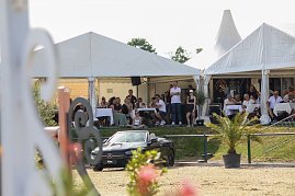 Zuschauer sitzen unter wei&szlig;en Eventzelten an Tischen und verfolgen ein Springreitturnier. Vor den Zelten steht ein schwarzes Mercedes-Benz Cabrio, dahinter bl&uuml;hende Pflanzen und ein sonniger Himmel.