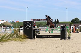 Ein Reiter in schwarzer Reitkleidung und Helm springt mit einem braunen Pferd &uuml;ber ein Hindernis mit Mercedes-Benz-Logo. Im Hintergrund sind Zuschauer unter einem wei&szlig;en Zelt, mehrere Fahnen und eine gr&uuml;ne Landschaft zu sehen.
