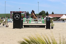Eine Reiterin springt mit einem braunen Pferd &uuml;ber ein Mercedes-Benz-Hindernis auf dem Sandplatz beim Reitturnier in Immenrode. Im Hintergrund sind weitere Hindernisse, gr&uuml;ne Landschaft und wei&szlig;e Zelte zu sehen.