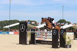 Ein Reiter springt mit einem braunen Pferd &uuml;ber ein Mercedes-Benz-Hindernis auf dem Sandplatz beim Reitturnier in Immenrode. Im Hintergrund sitzen Zuschauer unter wei&szlig;en Trib&uuml;nend&auml;chern, dar&uuml;ber wehen zahlreiche L&auml;nderflaggen.