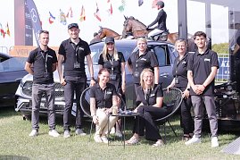 Gruppenfoto des Teams vom Autohaus Peter beim Reitturnier in Immenrode: Acht Teammitglieder in schwarzen Polohemden posieren vor ausgestellten Mercedes-Benz Fahrzeugen, im Hintergrund reiten Teilnehmer auf braunen Pferden &uuml;ber den Turnierplatz.