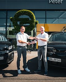 Vor dem Autohaus Peter stehen zwei Männer zwischen zwei schwarzen Opel-Fahrzeugen. Einer der Männer überreicht dem anderen einen Autoschlüssel, beide lächeln und geben sich die Hand. Im Hintergrund ist das Opel-Logo aus grünem Pflanzenmaterial an der Glasfassade zu sehen. Die Szene wirkt freundlich und sonnig.
