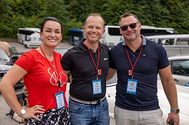 Christina Dost, Carsten Dost und Enrico Dost stehen Arm in Arm vor geparkten Fahrzeugen, tragen Namensschilder und rote Lanyards. Im Hintergrund sind Busse, Autos und gr&uuml;ne B&auml;ume zu sehen � die Szene bei der Rallye wirkt fr&ouml;hlich.