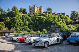 Reihe von klassischen Autos, darunter ein wei&szlig;er Renault 5 Turbo mit Startnummer 52, ein roter und ein cremefarbener Porsche, die auf einem Parkplatz vor einer gr&uuml;nen bewaldeten Anh&ouml;he stehen. Im Hintergrund thront das gelbe Schloss Hohenschwangau in Bayern unter blauem Himmel. Die Szene wirkt sonnig und freundlich.