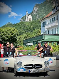 Das Team vom Autohaus Peter steht vor einem wei&szlig;en Mercedes-Benz Oldtimer mit Startnummer 15. Zwei Personen stehen links neben dem Auto, zwei rechts. Alle tragen schwarze Shirts, Sonnenbrillen und zeigen Daumen nach oben. Im Hintergrund ist Schloss Neuschwanstein auf einem bewaldeten Berg zu sehen, daneben ein wei&szlig;es Geb&auml;ude mit gr&uuml;nen Fensterl&auml;den. Sonniger, klarer Tag.