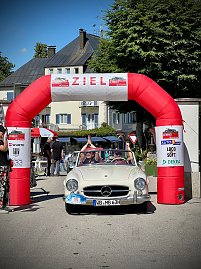 Wei&szlig;er Mercedes-Benz Oldtimer mit Startnummer 15 f&auml;hrt unter einem roten Zielbogen mit der Aufschrift �ZIEL� bei einer Rallye ein. Im Auto jubeln die Insassen mit erhobenen Armen. Menschen stehen rechts und links, klatschen und feiern. Im Hintergrund sind Geb&auml;ude mit Balkonen und B&auml;ume zu sehen. Sonniger Tag, festliche Stimmung.