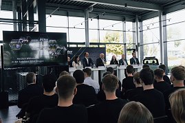 Podium mit sieben Mitgliedern der Gesch&auml;ftsf&uuml;hrung und Ausbildungsleitung der Autohaus-Peter-Gruppe bei der Begr&uuml;&szlig;ung der neuen Auszubildenden. Davor sitzen Azubis in schwarzen T-Shirts und h&ouml;ren zu.