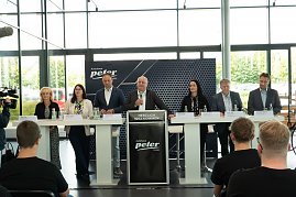 Podium mit sieben Mitgliedern der Gesch&auml;ftsf&uuml;hrung und Ausbildungsleitung der Autohaus-Peter-Gruppe bei der Begr&uuml;&szlig;ung der neuen Auszubildenden. Davor sitzen Azubis in schwarzen T-Shirts und h&ouml;ren zu.