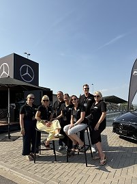 Gruppenfoto des Teams Peter beim Leichtathletikmeeting in Sondershausen. Sieben Personen tragen schwarze Polos mit Logo. Zwei sitzen auf hohen Barhockern, die anderen stehen daneben. Im Hintergrund sind ein schwarzer Mercedes-Cube mit großem Logo sowie ein schwarzer Mercedes CLA zu sehen. Alle lächeln und posieren fröhlich in der Sonne.