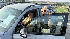 Ein Interessent sitzt am Steuer eines silbernen Mercedes GLB 220 4MATIC, während ihn eine Peter-Mitarbeiterin mit Sonnenbrille und schwarzem Polohemd freundlich berät. Auf der Windschutzscheibe ist ein Datenblatt mit Fahrzeugdetails sichtbar. Im Hintergrund ein Stadion mit Tribüne und grüner Rasen..