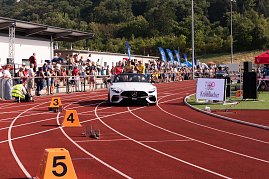Weißes Mercedes-Cabrio fährt auf die rote Tartanbahn des Stadions ein. Auf dem Rücksitz sitzen mehrere Sportlerinnen, während Zuschauer am Rand applaudieren.