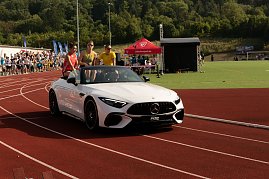 Nahaufnahme des weißen Mercedes-Cabrios auf der Laufbahn. Drei Sportstars sitzen auf der Rückbank, jubeln und winken dem Publikum zu.