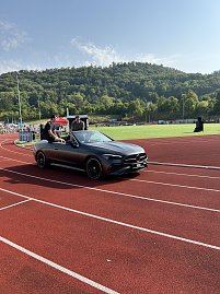 Graues Mercedes-Cabrio fährt auf die Tartanbahn ins Stadion ein. Zwei Personen sitzen oben auf der Rückbank. Im Hintergrund Zuschauertribünen und grüne Hügel.