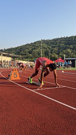 Sprinter im roten Trikot steht im Startblock auf Bahn 5, bereit für den Lauf. Weitere Athleten bereiten sich auf den benachbarten Bahnen vor. Zuschauer und Kampfrichter am Rand der Tartanbahn, im Hintergrund grüne Hügel.