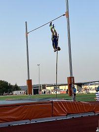 Stabhochspringer in blau-gelbem Trikot in der Luft kopfüber über der Latte. Im Hintergrund das Stadion und Zuschauer.