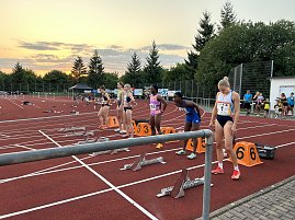 Sechs Sprinterinnen stehen an ihren Startblöcken auf der Laufbahn. Der Himmel im Hintergrund zeigt einen Sonnenuntergang mit Wolken.