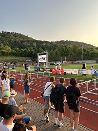 Zuschauer blicken von der Tribüne auf die Laufbahn. Eine große LED-Leinwand zeigt das Logo des Autohauses Peter. Im Hintergrund Waldhügel bei Sonnenuntergang.