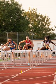 Drei Athletinnen im Hürdenlauf springen gleichzeitig über eine Hürde auf der roten Tartanbahn. Im Vordergrund sind die Hürden mit der Aufschrift ‚Sportzentrum am Göldner‘ zu sehen. Zuschauer und Kampfrichter im Hintergrund.
