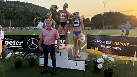 Drei Sportlerinnen stehen nach einem Leichtathletik-Wettkampf auf einem Siegerpodest und halten Blumensträuße in den Händen. Davor steht Centerleiter Marko Wuttke. Im Hintergrund sind eine grüne Sportanlage, Zuschauer sowie eine Waldkulisse bei Sonnenuntergang zu sehen. Vorne am Podest hängen Werbebanner, unter anderem vom Autohaus Peter und Mercedes-Benz.