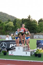 Drei Athletinnen stehen auf dem Siegerpodest, alle halten Blumensträuße. Die Siegerin in der Mitte trägt ein rotes Trikot, die beiden anderen blaue Outfits. Im Hintergrund das Stadion und grüne Hügel.