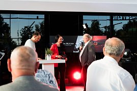 Christina Dost im roten Blazer spricht mit Helmut Peter im grauen Anzug. Beide stehen auf einer B&uuml;hne im Autohaus, links daneben, ebenfalls auf der B&uuml;hne, Andreas Peter. Das Banner He�s Mercedes � Cars, BBQ & Style ist im Hintergrund zu sehen.