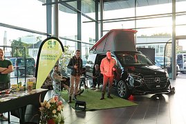 Ein schwarzer Mercedes-Camper mit ge&ouml;ffnetem Aufstelldach steht auf gr&uuml;nem Kunstrasen im Autohaus. Daneben zwei M&auml;nner, einer zeigt den Daumen hoch. Links im Bild ein Verkaufsstand mit Prospekten und Produkten.