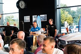 Zwei M&auml;nner stehen an einem kleinen Tisch, einer tr&auml;gt ein blaues Hemd mit DTZ-Logo, der andere ein schwarzes Polohemd. Sie pr&auml;sentieren Produkte, im Hintergrund sind ein schwarzer Mercedes und eine gro&szlig;e Uhr zu sehen.