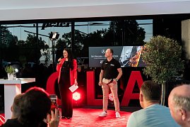 Christina Dost im roten Blazer spricht mit einem Mikrofon auf der B&uuml;hne. Neben ihr steht ein Mann im schwarzen Polohemd mit Autohaus-Logo. Hinter ihnen eine gro&szlig;e LED-Wand mit He�s Mercedes � Cars, BBQ & Style.