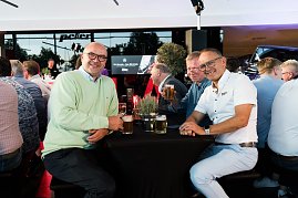 Drei M&auml;nner sitzen an einem Stehtisch mit Getr&auml;nken und lachen in die Kamera. Viele weitere G&auml;ste sitzen im Hintergrund an Tischen.