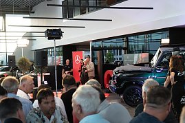 Blick in die Eventhalle mit vielen G&auml;sten im Vordergrund. Auf der B&uuml;hne stehen Christina Dost im roten Blazer und ein Mann in beiger Jacke, daneben ein schwarzer Mercedes-Gel&auml;ndewagen.