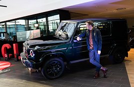 Ein junger Mann lehnt entspannt an einem schwarzen Mercedes-Gel&auml;ndewagen und schaut zur Seite. Der Wagen ist im Ausstellungsbereich mit Scheinwerfern beleuchtet.