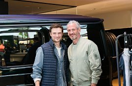 Zwei M&auml;nnermodels stehen nebeneinander vor einem schwarzen Mercedes. Beide l&auml;cheln in die Kamera, einer tr&auml;gt eine helle Bomberjacke, der andere eine dunkle Weste.