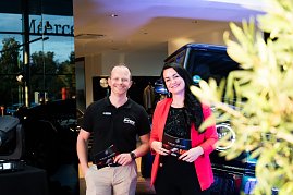 Eine Frau im roten Blazer (Christina Dost) und ein Mann im schwarzen Polohemd (Carsten Dost) stehen nebeneinander und l&auml;cheln. Beide halten Moderationskarten, im Hintergrund ein Mercedes-Gel&auml;ndewagen.