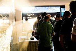 Eine Gruppe M&auml;nner steht in einer Schlange am Buffet. Sonnenlicht f&auml;llt durch die T&uuml;r, im Hintergrund ein Pavillon mit Autohaus-Peter-Logo.