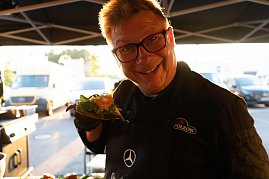 Ein Koch mit Brille und schwarzer Sch&uuml;rze mit Mercedes-Logo h&auml;lt l&auml;chelnd ein belegtes Sandwich in die Kamera. Im Hintergrund ein Foodtruck und Fahrzeuge im Sonnenlicht.