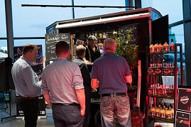Mehrere M&auml;nner stehen an einem Gin-Stand mit schwarzem Verkaufswagen. Auf Schildern steht Luzifer Gin und Gin-Cocktails. Regale mit Flaschen sind daneben aufgestellt.