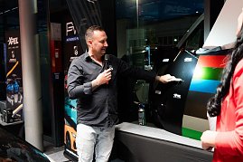 Ein Mann in schwarzem Hemd erkl&auml;rt mit Mikrofon verschiedene Lack- und Folienmuster an einer Autot&uuml;r. Daneben bunte Farbfolienstreifen.
