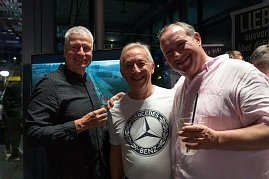 Drei M&auml;nner stehen nebeneinander und lachen in die Kamera. Einer tr&auml;gt ein wei&szlig;es Mercedes-Benz-T-Shirt, die anderen dunkle Hemden. Alle haben Getr&auml;nke in der Hand.
